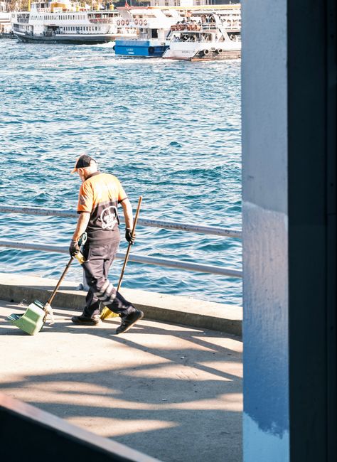 A worker from Docklands Carpet Cleaners dressed in an orange high-visibility shirt and dark pants, using a broom and dustpan to sweep debris along a paved walkway by a harbor. The background features 
