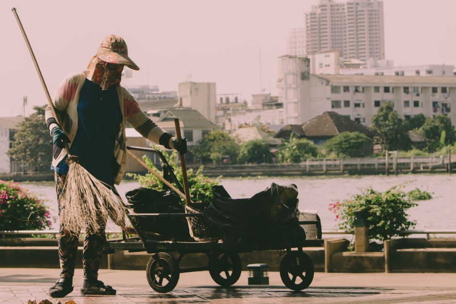 A worker from Docklands Carpet Cleaners dressed in an orange high-visibility shirt and dark pants, using a broom and dustpan to sweep debris along a paved walkway by a harbor. The background features 