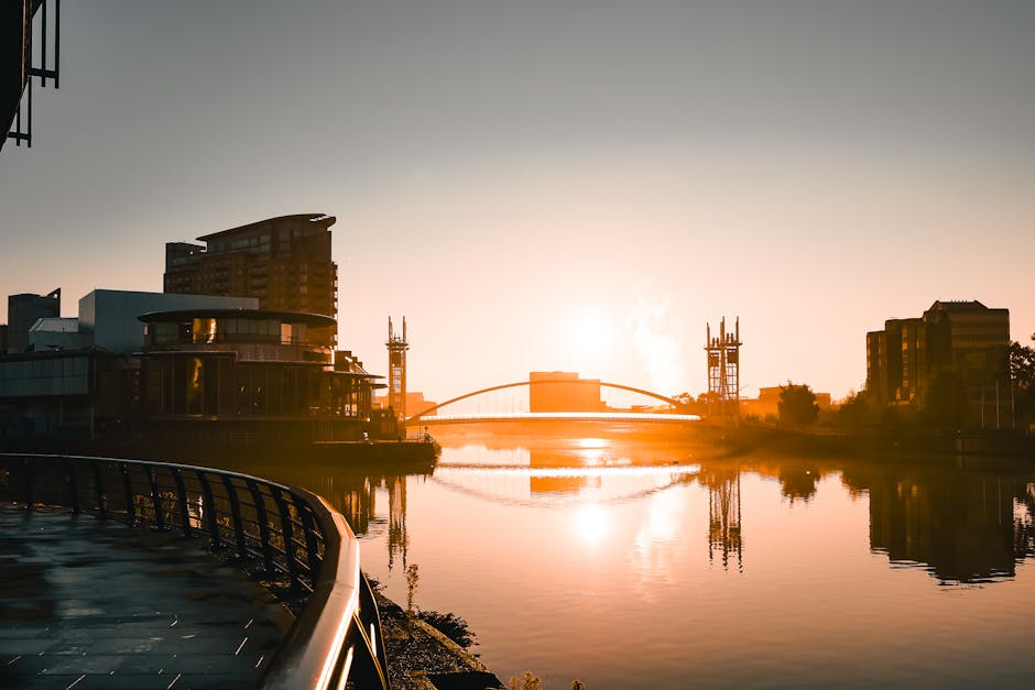 A daytime view of West India Quay in Docklands, showing modern commercial and residential buildings with reflective glass facades lining the calm waterway. The water surface is smooth with gentle ripp